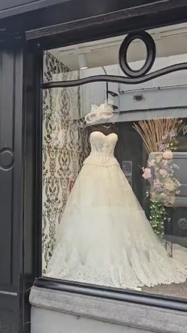 This video shows a display window of a clothing store, likely a bridal or formal wear boutique. The display features several mannequins adorned with dresses.

In the leftmost section of the display, a large, ornate white wedding dress with a strapless bodice and a voluminous lace skirt is prominently featured. A white hat with netting is placed on the mannequin's head. The background behind this dress is a textured wallpaper with a damask or floral pattern.

Moving to the right, the display transitions to feature two mannequins wearing elegant, floor-length teal or aqua-colored gowns. These dresses have draped bodices and flowing skirts. Accessories such as clutches and high-heeled shoes are placed at the base of these mannequins. A large floral arrangement with pink, purple, and white flowers and greenery is positioned between the wedding dress and the teal gowns.

Further to the right, another section of the display shows a mannequin in a white lace dress with long sleeves. This dress has a fitted silhouette and appears to be a different style of wedding or formal gown.

The far right of the display shows a mannequin wearing a blue and white patterned dress with a high neckline and a slit.

The video is shot from outside the store, looking through the glass window. Reflections of the street, including a parked vehicle, are visible on the glass, obscuring parts of the display at times. The lighting in the display window appears to be artificial, highlighting the garments. The overall impression is of a well-arranged and sophisticated display of formal attire.