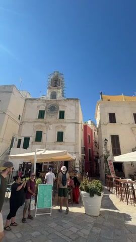 The video pans across an outdoor square with people walking and sitting at tables. Several buildings line the square, including a multi-story structure with a clock tower and bell tower, and another building with a white facade and green shutters. Outdoor seating areas with umbrellas and tables are set up in front of some buildings, suggesting a cafe or restaurant. In the center of the square, a large white planter holds greenery. In another area of the square, rows of blue chairs are set up facing a stage with a large banner. The sky is clear and blue, indicating a sunny day. People are dressed in casual summer clothing.
