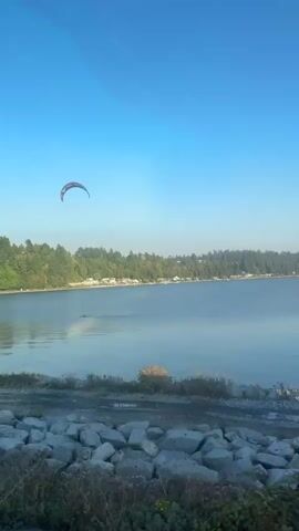 A wide shot shows a body of water with a shoreline covered in trees and houses in the background. In the sky, a red and black kite is visible. Below the kite, a person is on the water, appearing to be kiteboarding. The water is calm, with gentle ripples. The sky is a clear blue. Rocks and some dry vegetation are in the foreground.