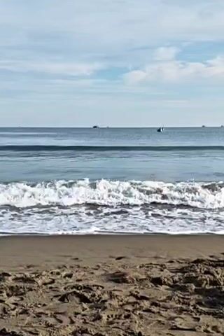 The video shows a beach scene with gentle waves lapping at the shore. The sand in the foreground is textured with footprints. In the background, the calm ocean stretches to the horizon, dotted with a few distant boats. The sky above is partly cloudy with a pale blue hue. The camera is static, focusing on the interplay between the sand, sea, and sky.