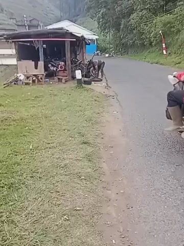 A scene is captured from the perspective of someone walking or riding, showing a rural roadside environment. On the left, a small, open-air structure serves as a makeshift workshop or repair stall. Inside and around it are several motorcycles and what appears to be a red air compressor. A person is bent over, seemingly working on one of the motorcycles. To the right of the structure, a dirt path borders a grassy slope. The main focus shifts to the paved road where a motorcycle carrying three people is traveling away from the viewer. The rider is wearing a red shirt and a dark helmet. The two passengers are also on the motorcycle; one is wearing a blue hijab and a pink top, and the other is wearing a dark top. Further down the road, another motorcycle is visible, also moving away from the viewer. The background shows a verdant hillside with some structures and trees, suggesting a mountainous or hilly rural area. The lighting indicates it is daytime, possibly overcast.
