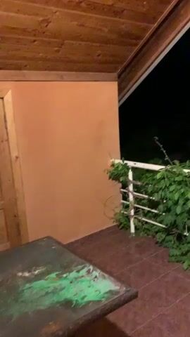 The video shows a balcony at night. There is a wooden ceiling and an orange wall. A white railing is covered in green vines. A table with a green and white surface is visible in the foreground. In the background, there is darkness with some trees and a distant structure with lights. The camera pans across the scene.