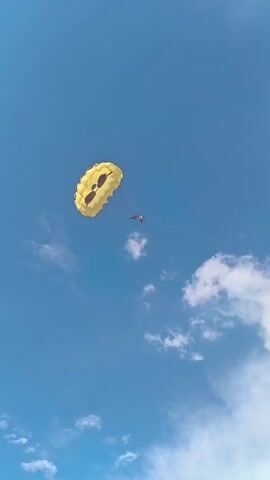 A yellow parachute with dark accents is suspended against a bright blue sky with scattered white clouds. A person or two are visible in a harness suspended beneath the parachute, appearing to be parasailing. The image is taken from a low angle looking up at the parachute. The scene suggests a daytime activity, likely in a warm weather location, possibly near water given the nature of parasailing. There is no visible text in the image.