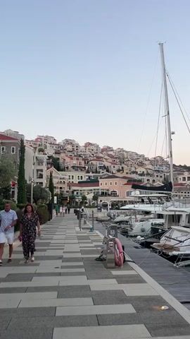 The video shows a person walking along a paved walkway next to a marina. The walkway is made of gray and light gray rectangular tiles. On the left side of the walkway are buildings and landscaping. On the right side of the walkway are boats docked in the marina. In the background, there are more buildings on a hill. The sky is clear and the light suggests it is either early morning or late afternoon. The person walking in the foreground is a woman wearing a dark floral print dress.