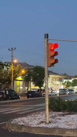 The video shows a street scene at dusk. A traffic light is visible in the foreground, showing a red light. Cars are waiting at the light. In the background, there are buildings, trees, and streetlights. The sky is a clear blue. Some cars appear to be emitting exhaust fumes.