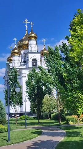 This is a vertical video of an Eastern Orthodox church with golden domes on a sunny day. The church is white with multiple domes, each topped with a cross. The building is surrounded by lush green trees and manicured lawns, with a paved walkway leading towards it. The sky is a vibrant blue with a few white clouds. The scene appears peaceful and serene, likely indicating a daytime setting in a temperate climate. There is no visible text in the video. The location context provided is Bryukhovetskaya, Russia.