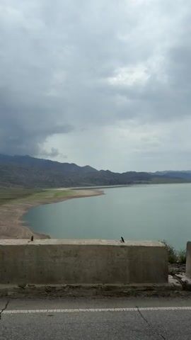 The video shows a wide shot of a large body of water, likely a reservoir, with mountains in the background under a cloudy sky. In the foreground, there is a concrete barrier or wall above a road. The water is a greenish-blue color and appears calm. The shoreline in the distance is visible, showing exposed earth and some vegetation. There are no people or animals visible in the frame. The lighting suggests it is daytime, possibly overcast.