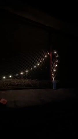 The video opens with a shot of a dark outdoor area at night, illuminated by a string of small, warm-toned lights draped across a mesh fence and a red pole. A white object, possibly a box or container, is visible at the base of the pole. The camera then pans upwards and to the right, showing the silhouette of trees against a dark blue twilight sky. The rest of the video continues to show dark silhouettes of trees and a railing against the darkening sky, with no further details visible.