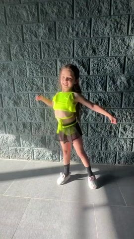 A young girl, likely a child, is dancing in front of a grey, textured brick wall. She is wearing a neon yellow crop top with ruffled sleeves and a matching neon yellow string tied around her waist. Her lower half is clad in a black mesh skirt with a neon yellow trim. She is also wearing white sneakers with black accents and grey socks. Her hair is dark and pulled back, and she has a wide smile on her face, appearing joyful and energetic as she dances. Her arms are in motion, suggesting a lively dance routine.

The setting appears to be indoors, possibly a studio or a room with a modern aesthetic. The floor is tiled with large grey squares. Sunlight is casting shadows on the floor, indicating it is daytime. The overall mood is playful and cheerful, driven by the girl's enthusiastic performance. There is no visible text in the media file. The location context of Sverdlovskiy, Russia, does not provide specific visual cues within the frame.