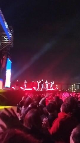 The video captures an outdoor nighttime concert or festival from within the crowd. Numerous people are visible, some with their hands raised in the air, indicating engagement and excitement. The scene is illuminated by vibrant stage lighting, with prominent red beams shining across the stage and audience, alongside some blue light from screens. A stage is visible in the distance, featuring performers, though they are small and indistinct. Large screens on either side of the stage display visuals. The atmosphere appears lively, with the crowd enjoying the performance. The video is oriented vertically.