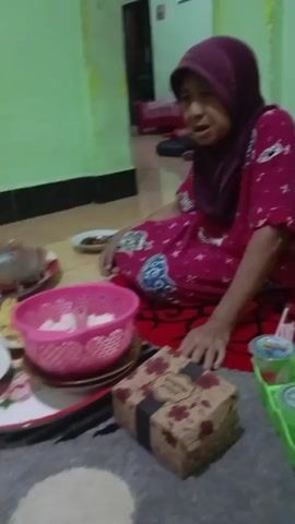 The video shows an elderly woman wearing a headscarf and a pink patterned dress, sitting on the floor. She is looking at something to her right with a concerned expression. In front of her is a box wrapped in brown paper with a black ribbon and floral designs. To her left is a pink basket filled with white rice, and to her right is a green plastic tray holding several small cups with lids and straws. The environment appears to be an indoor setting with green walls and a tiled floor. The lighting is dim.