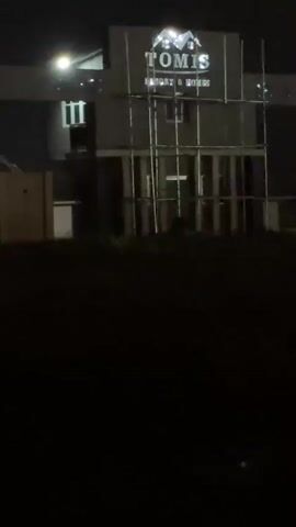 The video is taken at night and focuses on a building with the sign "TOMIS RESORT & HOMES" displayed on the front. Scaffolding surrounds part of the building's facade. The camera pans slightly to the right and then down, eventually focusing on a dark, unidentifiable ground area. In the background, a faint light source is visible on the horizon.