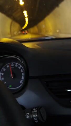 The video, shot from inside a car, shows the vehicle driving through a tunnel at a moderate speed, as indicated by the speedometer on the bottom left. The tunnel is well-lit with rows of lights along the ceiling, and its walls are a combination of gray and white sections, some with arch patterns. A few other cars are visible in the distance, ahead of the camera. The windshield of the car appears slightly speckled, possibly with dust or water droplets. The overall impression is of a routine drive through a modern tunnel.