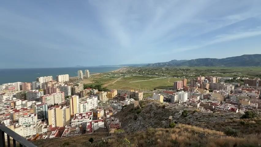 This is a wide-angle, panning video shot from a high vantage point overlooking a coastal city. The foreground is dominated by a rocky, dry hillside covered in sparse, dry grass. Below the hill, a dense urban area stretches out, characterized by numerous multi-story buildings with light-colored facades, some appearing residential and others more commercial. Beyond the city, a long, sandy beach meets the blue ocean. Further inland, a flat, green plain is visible, bordered by distant, hazy mountains under a bright blue sky with scattered white clouds. The camera pans slowly from left to right, offering a panoramic view of the urban landscape, coastline, and natural surroundings. There are no people or discernible actions visible in the video. The lighting suggests it is daytime, possibly late afternoon or early morning given the soft light.