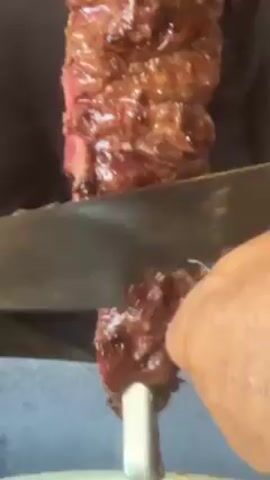 Here is a description of the video:

A person is cutting meat off a skewer with a knife. The meat appears to be beef, glazed and cooked to a medium-rare doneness. The skewer is stainless steel.  The person is seated in what looks like a restaurant. The background is softly blurred.