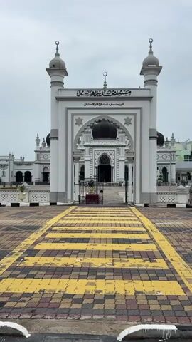 The video shows a wide shot of the exterior of a mosque. The mosque is white with a large arched entranceway leading to the main building. The entranceway is flanked by two towers, each topped with a dome. Above the archway, there is an Arabic inscription. The ground in front of the mosque is paved with colorful tiles in a checkerboard pattern, with yellow and black stripes creating a walkway. A silver van enters the frame from the left and drives across the tiled area in front of the mosque before stopping. The sky is overcast.