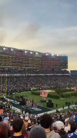A wide shot reveals a packed stadium during what appears to be a halftime show. People fill the stands, while on the field, a stage area decorated with green landscaping is the focal point. Performers, possibly musicians, are gathered on the stage. The massive screen above the stadium reads "Levi's Stadium" and displays video of the event happening on the field. The sky overhead is cloudy, with a hint of sunset colors.