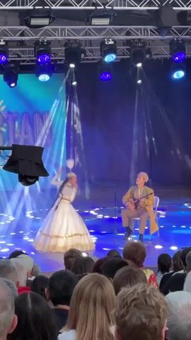 A live performance is taking place on an outdoor stage. A large banner displaying the word "STAN" with a sun-like graphic and what appears to be a stylized eagle is visible behind the performers. The stage is illuminated with bright blue and white lights, creating a vibrant atmosphere. In the foreground, the back of an audience's heads is visible, indicating they are seated and watching the performance. On the stage, a female dancer in a traditional white and gold dress is gracefully moving, her arms extended and her skirt swirling. She appears to be in the midst of a pirouette or a similar dynamic dance move. To her right, a male musician is seated on a chair, playing a stringed instrument that resembles a mandolin or lute. He is dressed in traditional attire, including a light-colored jacket over a darker garment and a head covering. The overall scene suggests a cultural performance, possibly a celebration or festival, with a focus on traditional music and dance.