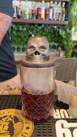 The video shows a close-up shot of a cocktail shaker with a skull-shaped lid, filled with a dark red liquid and ice. The shaker is placed on a bar counter with other bar items visible in the background. A person's arm and shoulder are visible on the left side of the frame. The scene appears to be in a bar or restaurant setting with shelves of bottles and greenery in the background. The lighting is warm and inviting. The overall impression is that of a unique and possibly spooky cocktail being prepared.