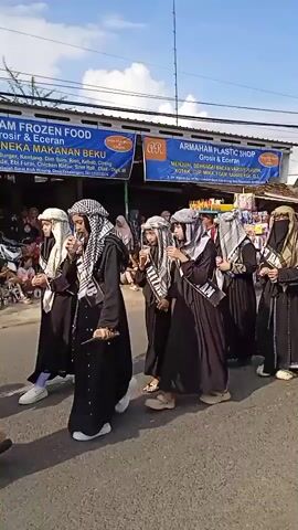 A group of people are walking in a parade or procession on a paved road. 
In the foreground, a line of people are dressed in long black robes and headscarves resembling traditional Arabic attire. Some of the figures wear full face coverings. 
Behind them, another group follows, wearing black long-sleeved shirts, batik-patterned skirts, white sneakers, and headpieces. Most of them wear dark headscarves.
The setting is an outdoor environment during daytime with blue skies. A building with blue banners stands beside the road, advertising "Frozen Food" and a "Plastic Shop." Additional onlookers and some parked motorcycles are visible in the background.
