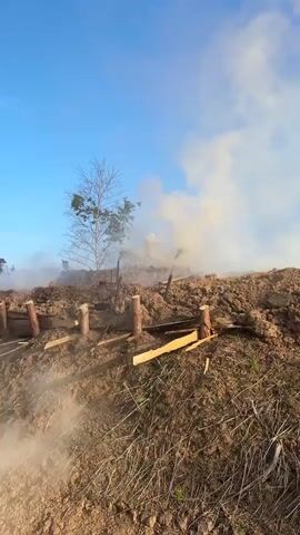 A video taken from a low angle shows a landscape with a clear blue sky and some clouds. In the foreground, there is dry grass and brown soil. A rudimentary barrier made of wooden posts and planks is visible. Behind this barrier, the ground rises, covered in brown soil and debris. A tree with no leaves stands on the left side of the frame. Smoke is rising from behind the hill, obscuring part of the sky and a few wooden structures. The smoke appears to be originating from a fire.