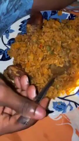 A person with dark skin is eating a meal off a plate. The plate has a blue and white floral pattern. The meal consists of orange-colored rice, with visible bits of green and orange vegetables, and two pieces of dark brown fried food. The person is using a silver fork to take a bite of the rice. The plate is sitting on an orange and white patterned surface. The person is wearing a blue and white shirt. The person is holding a cell phone in their other hand.