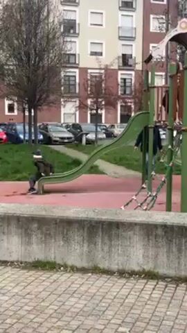 A child in a dark jacket and light-colored hat is running away from a green playground slide on a red surface. A man in a dark jacket is walking toward the playground equipment, which has climbing ropes. The playground is in front of a multi-story building with many windows and a few trees with bare branches. Cars are parked along the curb beyond the playground. The camera is behind a low concrete wall with brick pavers in the foreground. The sky is overcast.