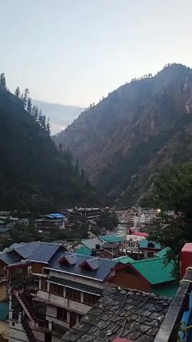 The video presents a panoramic view of a bustling mountain town nestled within a deep, verdant valley. The camera slowly pans from left to right, revealing the expansive landscape and the community built into it.

Steep, heavily forested mountainsides, densely covered with coniferous trees, flank both sides of the valley, their upper reaches fading into an overcast, diffused sky. Below, a wide, rocky riverbed with a flowing river cuts through the heart of the town. The water appears light blue-green in some sections.

Numerous multi-story buildings, characterized by varied architectural styles, line the riverbanks and climb the lower slopes of the valley. Many feature distinct pitched roofs in vibrant shades of teal-green, red, dark gray, and blue. Building facades are predominantly light-colored – beige, cream, or white, with some exposed stone or brickwork. Balconies and external staircases are common.

As the camera continues its pan and slightly zooms in, a large, densely packed parking lot comes into view, filled with numerous cars, mostly white and yellow taxis, and several buses, indicating a busy area. Small figures, possibly people, can be seen on a path or street near the parking area. In the immediate foreground, power lines and utility structures are visible on rooftops.

The scene depicts an active mountain community, likely a tourist or pilgrimage destination like Manikaran, India. The soft, even lighting and lack of harsh shadows suggest either early morning or late afternoon, with calm, overcast weather. No discernible text is visible in the video.