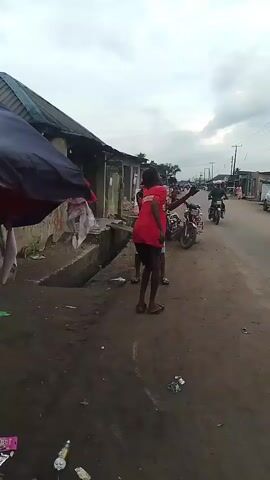 On a cloudy day, a person in a red t-shirt and dark shorts stands on a dirt road, facing the camera. They are gesturing with their arms outstretched to the sides. A second person, dressed in a red shirt and blue shorts, stands next to them to the left. A gutter runs alongside the road, and a dilapidated building is visible behind them, partially covered by a blue and black awning. Motorbikes and a white truck are driving on the paved road in the background, with several buildings and utility poles lining the roadside. Litter is scattered on the ground.