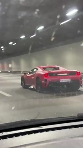 A car drives down a highway in a tunnel. The car in the foreground filming through the windshield is behind and following a red Ferrari, which is in the middle lane. The tunnel lights are bright, and there are direction signs overhead indicating Zurich City and Basel.