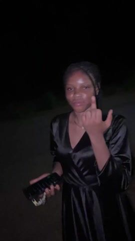 A young, dark-skinned woman is standing outside at night. She is wearing a shiny black dress with a v-neck and long sleeves. Her hair is braided back, and she has a nose piercing. The woman is holding a black smartphone in her left hand. In the video, the woman sticks her tongue out and touches her finger with it. She then wipes her finger in her mouth. The background is dark and blurry, suggesting an outdoor setting with minimal lighting.