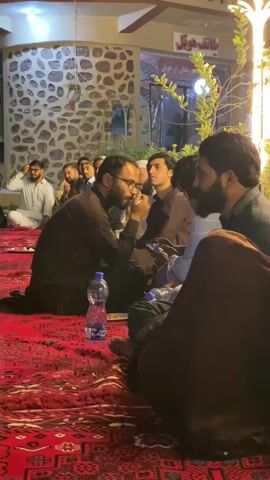 The video shows a gathering of men sitting on a red carpet in an outdoor setting. The men are dressed in traditional clothing. Some of them are holding or looking at their phones. There are bottles of water and food on the carpet. In the background, there is a building with patterned stone walls and a sign with writing in a foreign language. The scene appears to be a social or religious gathering.
