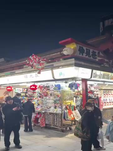 The video shows a bustling street scene at night. Several shops line the street, adorned with decorative red lanterns and other ornaments. People are walking, browsing, and taking pictures near the shops. The shops are lit up with bright lights, showcasing various goods on display. The overall environment appears to be a lively marketplace or shopping street in an urban area.