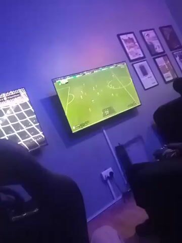 A first-person shot in a dimly lit room reveals someone playing a soccer video game on a wall-mounted TV, accompanied by background music. The game shows players in white jerseys and the field of play. In the foreground, the arm and leg of a person are visible, implying they are seated.  Another TV screen can be partially seen to the left, while framed pictures decorate the wall above the primary TV.  A small heater sits below the TV, along with a power outlet and cords. The room is lit with purple and blue light, creating a cool, subdued atmosphere.