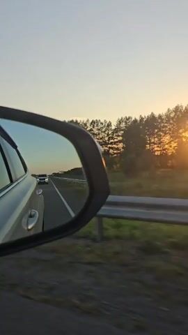 The video is taken from inside a moving car, looking at the passenger side mirror. The mirror reflects a highway with a white car driving in front of it. On the right side of the frame, there is a guardrail and a field of grass. Beyond the field, a line of trees is visible against a clear sky, with the sun setting behind them, casting an orange glow. The car appears to be traveling on a paved road, likely in a rural area, during daylight hours with the sun nearing the horizon.