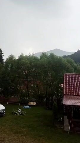 The video shows a yard with a house and a view of mountains. In the foreground, there is a red-roofed house with wooden accents and a porch. To the left of the house, there is a green lawn with some garden decorations, including a white swan figure and a blue object. A white car is partially visible on the left edge of the frame. In the background, there are lush green trees and rolling hills that lead up to mountains in the distance. The sky is overcast, suggesting a cloudy or possibly rainy day. The overall scene conveys a peaceful, rural atmosphere, likely in a mountainous region. The location is identified as Synevyrska Poliana, Ukraine, which is known for its natural beauty and mountainous terrain. No people are visible in the video. There is no discernible text in the video.