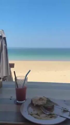 The video shows a view from a restaurant or similar establishment overlooking a sandy beach and a blue ocean under a clear blue sky. In the foreground, on a striped table, there is a glass of pink liquid with two straws, and a white plate with the remains of a meal consisting of what appears to be avocado toast topped with a poached egg. There is a fork and knife on the plate, indicating that the meal is partially consumed. The beach is mostly empty except for a few people swimming in the water in the distance. The scene is bright and peaceful.