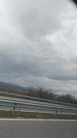 The video depicts a highway scene under a cloudy sky. A white delivery van and a white tanker truck pass by on the road, separated by a metal barrier. Trees with bare branches stand to the side of the highway.