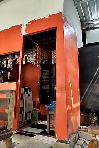 The video opens on a small outdoor market stall with an orange painted exterior. Shelves inside are stocked with various packaged goods, likely snacks or cigarettes. In front of the stall, a wooden counter is visible. The camera pans to the right, revealing a wooden table and benches, with a cat lying on one of the benches. The camera then pans further, showing a motorcycle parked next to a bicycle. A shaded canopy is overhead, with a black mesh netting displaying text, possibly advertisements or shop names. The overall environment appears to be an open-air, informal marketplace.