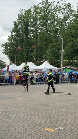 Three figures wearing black-and-neon-green outfits marked “15”, “23”, and “7” stand in a paved outdoor space before a crowd separated by a metal railing. Tents and trees can be seen beyond the crowd. The figure wearing “23” is on a tall bike. The figure wearing “15” holds a bat and raises his left arm. The figure wearing “7” stands with his feet apart. A fire hose and hula hoop lie on the pavement.