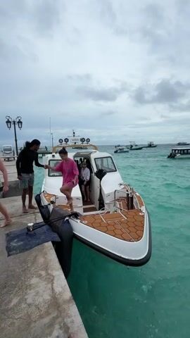 Here's a detailed description of the video:

The video showcases a vibrant scene at a harbor or marina. The foreground features a white boat docked at a pier. A man with dark hair is seen helping a woman in a pink outfit step off the boat onto the pier. A second woman wearing a white shirt and black shorts stands in the boat's doorway.

The boat has a brown, textured deck and a closed cabin. A Garmin device and multiple lights are visible on its roof. The water surrounding the boat is a stunning turquoise color.

The background reveals several other boats anchored in the water. Further out, a beach lined with palm trees is visible, and people can be seen on the sand. 

The sky is overcast with grey clouds, giving the scene a somewhat muted yet still visually appealing atmosphere. The overall impression is of a tropical or island setting.