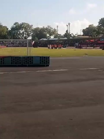 The video shows a soccer field from a distance. There are players in orange uniforms standing on the field. There are also spectators in the stands. A soccer net and some equipment are visible in the foreground.