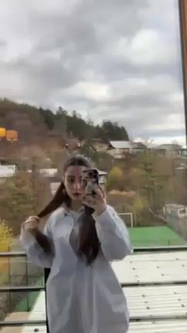 A young woman is standing on a balcony, taking a selfie with her phone. She is wearing a light blue oversized shirt over a t-shirt. Her long brown hair is parted in the middle and pulled back into two low ponytails. She is standing in front of a glass railing. Beyond the railing, there is a hillside with trees and buildings. In the foreground is a green field with a soccer goal. The sky is cloudy. The woman's phone has a case with images on it. She poses for the selfie.
