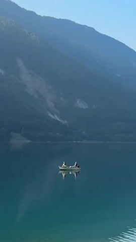 Two people in a small boat are fishing on a calm lake. The boat is white with a red stripe and is equipped with an outboard motor at the stern. One person is seated in the front of the boat, facing forward, while the other is seated in the back, facing backward, holding a fishing rod. The water is clear and reflects the boat and the people. Behind the lake, a steep, forested mountain slope rises, with patches of bare rock and a landslide scar visible. The sky is a pale blue, indicating daylight.