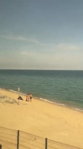 The video shows a sunny beach scene in Cabrera de Mar, Spain, as viewed from a moving perspective, likely a train or car. The camera pans along a golden sandy beach bordered by the calm, turquoise-blue Mediterranean Sea under a bright sky with scattered clouds.

Several people are visible on the beach. At the beginning, a small group of three people stands near a white sign on a post, and another person sits further down the shore. As the view progresses, other individuals and small groups are seen lounging, sitting on rocks, or lying on the sand. A line of rocks separates the sand from a slightly elevated paved area, and a metal fence runs along the immediate foreground. Towards the end of the clip, a large, light-colored metal structure, resembling a ladder or part of a signal post, briefly enters the frame, partially obscuring the view. Reflections of buildings can be seen in the window through which the video is filmed. The overall impression is one of a peaceful, leisurely day at the coast.