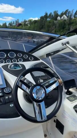 The video begins with a close-up view of a boat's steering wheel and dashboard, featuring various gauges and a compass. The perspective then shifts to show a marina with numerous boats docked under a bright blue sky with scattered clouds. In the distance, lush green hills dotted with houses form the backdrop. A white speedboat with two people on board is seen navigating through the marina. The camera pans across more docked sailboats and motorboats, highlighting the serene waterfront environment. The video concludes with a wider shot of the marina, emphasizing the picturesque setting and the collection of vessels.