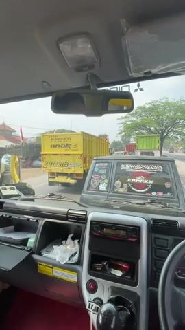 The video is a first-person perspective from inside the cabin of a truck. The dashboard and steering wheel are visible in the foreground. Through the windshield, the road and other vehicles are visible. A yellow truck with the words "BANGKIT ANAK SELEBRITIS" printed on its side is prominent in the middle distance. The sky is visible and appears to be overcast. The truck appears to be stationary, possibly in traffic.