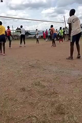 The video shows a group of young people, primarily men, playing volleyball on an outdoor dirt court in Ngenyilel ward, Kenya. The sky is overcast with clouds, indicating daytime. A white volleyball net stretches across the court, supported by poles. Players are actively engaged in the game, with one person in a yellow shirt serving or hitting the ball over the net towards the opposing team. Other players are positioned on both sides of the net, ready to receive or return the ball. The ground is mostly bare dirt with patches of dry grass. One player in a light-colored shirt on the right side of the frame has the word "CHRIST" visible on the back of their shirt.