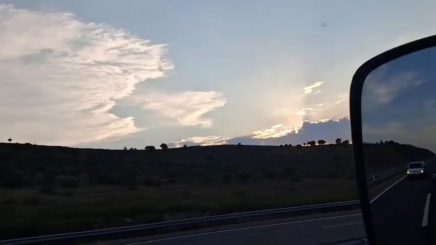 A video shows a view from a vehicle, likely a truck or van, looking out a side mirror. The mirror reflects a dark silhouette of the vehicle's side. The main scene is a landscape with a road in the foreground, bordered by a guardrail. A white van is visible driving on the road. Beyond the road is a silhouetted hill with scattered trees. The sky is a mix of blue and pink clouds, with the sun partially obscured by clouds, casting rays of light. The overall scene appears to be at dusk or dawn.