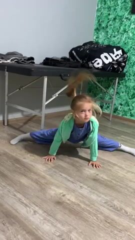A young girl with blonde hair is on the floor doing splits. She is wearing a light blue jumpsuit and a green hoodie. Her feet are white socks. The floor is wooden. Behind her is a black massage table with some items on it. There is a textured green wall in the background. The girl is looking at the camera. She then transitions into a kneeling position and waves her hands.

The video appears to be filmed indoors in a room that could be a dance studio or a multipurpose room. The lighting suggests it is daytime. There is text on a black bag on the table, but it is blurry and unreadable.